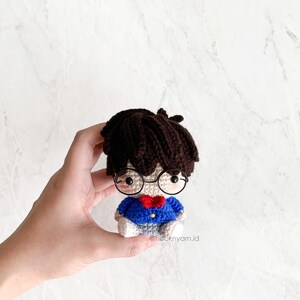 May include: A crocheted amigurumi doll with brown hair, wearing blue and white clothing, glasses, and a red bow tie. The doll is being held in a hand.