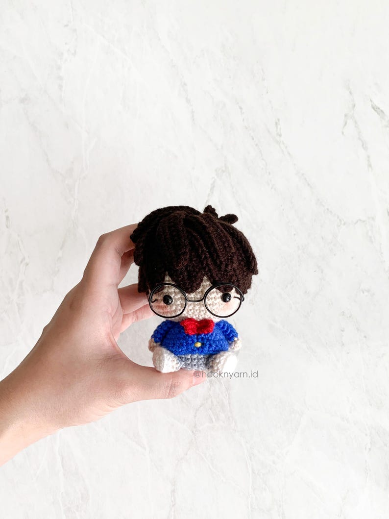 May include: A crocheted amigurumi doll with brown hair, wearing blue and white clothing, glasses, and a red bow tie. The doll is being held in a hand.