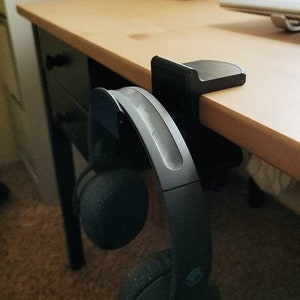 May include: Black headphone holder attached to a desk with a pair of black headphones hanging from it.