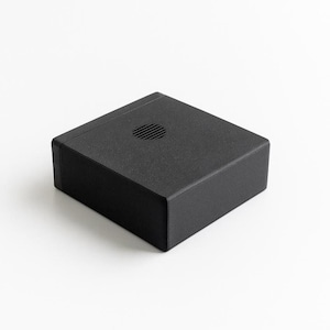 Könnte beinhalten: Eine matte, schwarze, rechteckige Box mit strukturierter Oberfläche. Ein kleines, kreisförmiges Lüftungsloch befindet sich mittig auf der Oberseite. Die Box steht vor einem weißen Hintergrund, der ihr schlichtes, modernes Design hervorhebt. Wahrscheinlich ein Gehäuse für Elektronik.