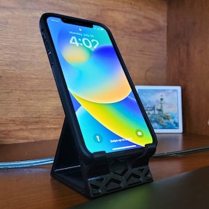 Pode incluir: Uma capa de telefone preta com um telefone dentro. O telefone está exibindo um fundo abstrato colorido com a hora 4:02. O telefone está sentado em um suporte de telefone impresso em 3D preto.