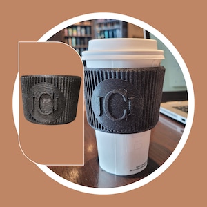May include: A black and white coffee cup sleeve with the initials "JCI" printed on it. The sleeve is made of a textured material and is designed to fit a standard-sized coffee cup.