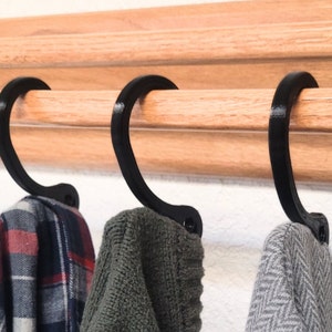 Peut inclure: Trois crochets en plastique noir sont montés sur une barre en bois. Les crochets tiennent une chemise à carreaux, un pull vert et un pull gris.