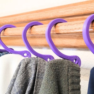 May include: Five purple plastic hangers with rounded edges hang on a wooden rack.  A blue denim jacket, a white shirt, a gray sweater, a brown sweater, and a black sweater are hanging on the hangers.
