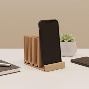 May include: A beige wooden phone stand with a black smartphone in a case. The stand has a unique, curved design with multiple slots. A small succulent in a white geometric pot is visible in the background.