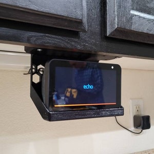 Può includere: Un display intelligente Echo Show nero montato sotto un pensile da cucina con una staffa in metallo nero. Lo schermo mostra la parola "echo" su uno sfondo blu.