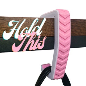 May include: Pink plastic hook with a chevron pattern, designed to hang items on a shelf or table edge. The hook is attached to a wooden surface and the text "Hold This" is visible in the background.