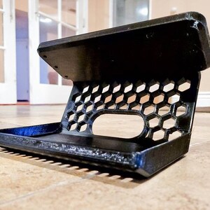 May include: A black 3D printed phone stand with a honeycomb design and a raised platform. The stand is designed to hold a phone in a vertical position.