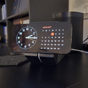 May include: A black digital clock displaying the time and the month of January. The clock has a circular analog clock face and a calendar. A black speaker is in the background. The clock is on a black stand.