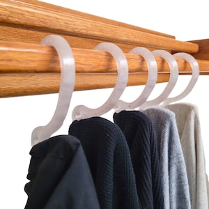 May include: Five pieces of clothing hanging on a wooden rack with clear plastic hooks. The clothing includes a black jacket, two black sweaters, a gray sweater, and a white sweater.