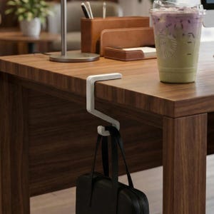 May include: A white, S-shaped desk hook is attached to the edge of a dark brown wooden desk. A black briefcase hangs from the hook. A cup of iced beverage sits on the desk, along with office supplies.