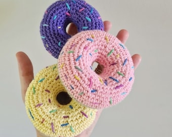 May include: Three crocheted doughnuts in yellow, pink, and purple with colourful sprinkles. The doughnuts are handmade and have a soft, textured surface.