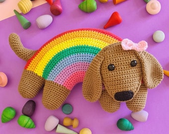 May include: A crocheted rainbow dachshund toy with brown fur and a pink bow. The toy is lying on a purple surface with wooden toys scattered around it.