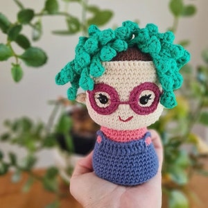 Puede incluir: Una muñeca de crochet con pelo verde, gafas rosas y un vestido de peto azul.
