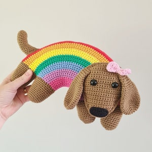 May include: A crocheted rainbow dachshund with a pink bow. The dachshund is brown and has black eyes. The rainbow is made of red, orange, yellow, green, blue, indigo, and violet yarn.