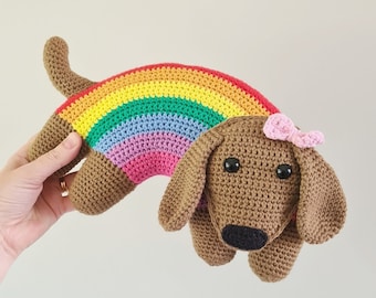 Puede incluir: Un dachshund de crochet arcoíris con un lazo rosa. El dachshund es marrón y tiene ojos negros. El arcoíris está hecho de hilo rojo, naranja, amarillo, verde, azul, índigo y violeta.