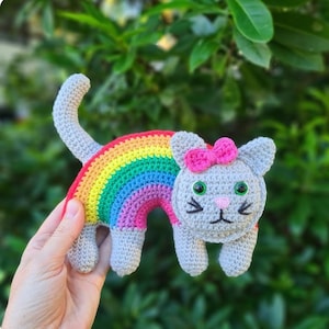 Op de afbeelding: Een grijs gehaakt katten speelgoed met een regenboogkleurige gehaakte staart. De kat heeft een roze strik en groene ogen.