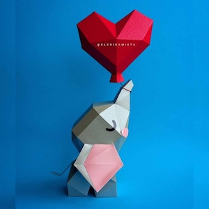 Può includere: Un elefante geometrico di carta grigio tiene in mano un palloncino rosso a forma di cuore su sfondo blu. L'elefante ha un bavaglino rosa e un piccolo occhio nero curvo. Il palloncino è attaccato con uno spago rosso. Il testo "@ELORIGAMISTA" è sul palloncino.