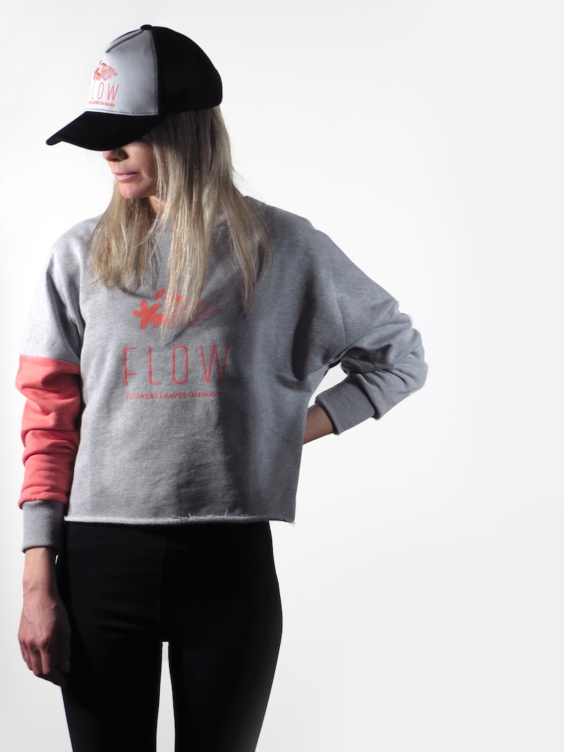 May include: A gray sweatshirt with a red graphic of a flower and the word "FLOW" with the text "FLOWERS LEAVE THEIR WAY" underneath. The sweatshirt has a red and gray color block design on the sleeves. The model is wearing a black and white baseball cap.