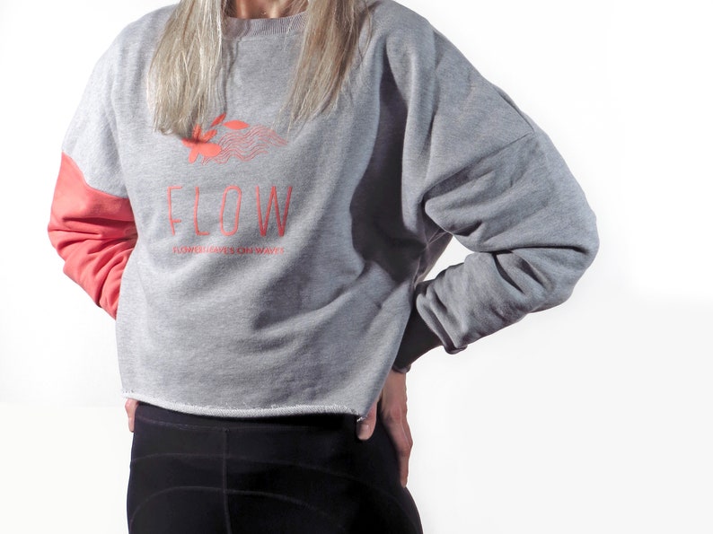 May include: A gray and coral cropped sweatshirt with the word "FLOW" and the text "FLOWERS GROW ON WAVES" printed on the front.