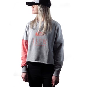 May include: A woman wearing a gray and pink cropped sweatshirt with the text "FLOW" and a black and white trucker hat. The sweatshirt has a flower graphic and the text "FLOWERS GROW WHEREVER THEY ARE PLANTED".