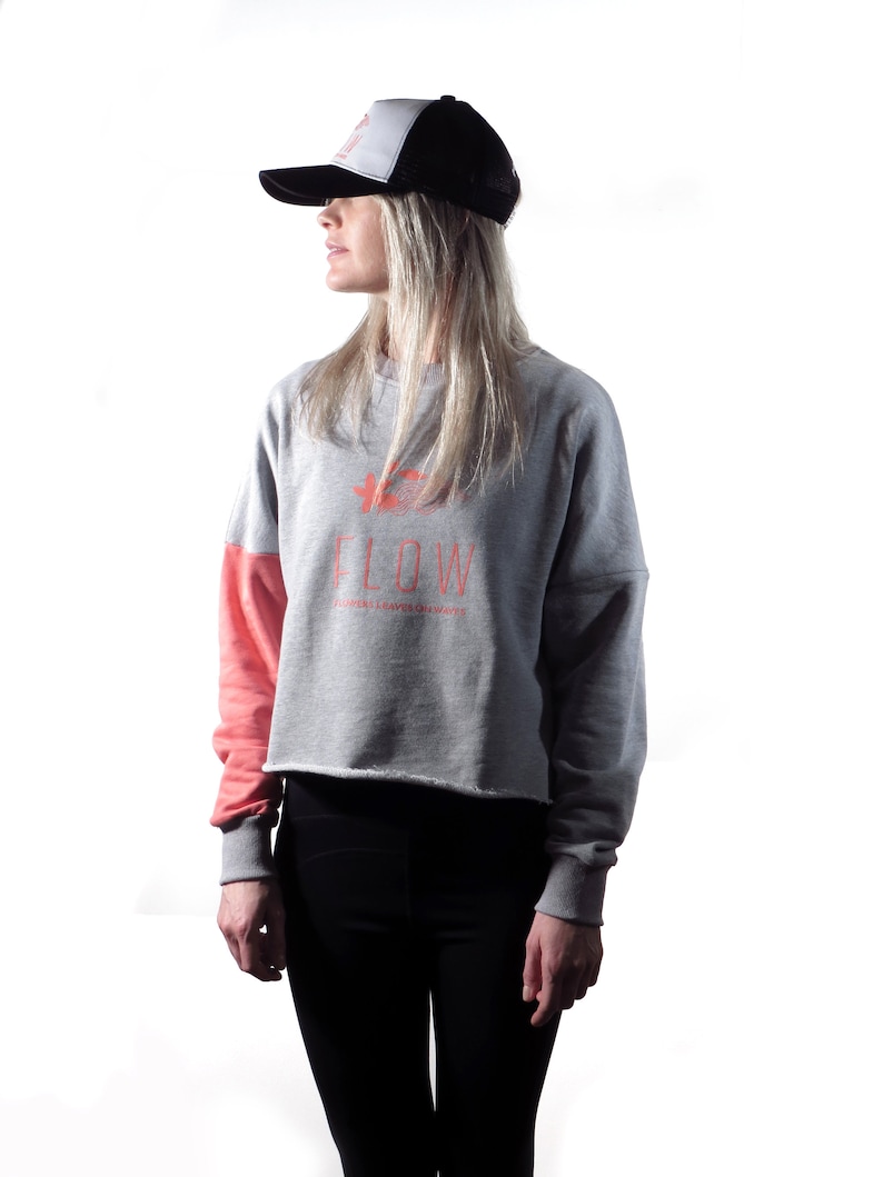 May include: A woman wearing a gray and pink cropped sweatshirt with the text "FLOW" and a black and white trucker hat. The sweatshirt has a flower graphic and the text "FLOWERS GROW WHEREVER THEY ARE PLANTED".