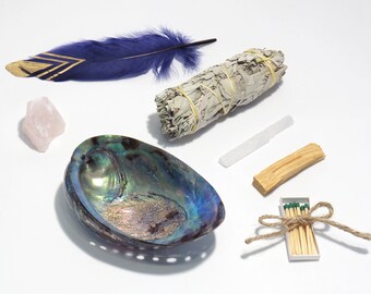 May include: A smudging kit with a white sage bundle, a palo santo stick, a clear quartz crystal, a pink rose quartz crystal, a blue and gold feather, a abalone shell, and a box of matches.