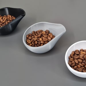 Puede incluir: Tres cucharas de café de cerámica en diferentes colores: negro, gris claro y blanco. Cada cuchara está llena de granos de café tostados.