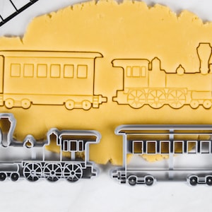 Op de afbeelding: Twee koekjesvormen in de vorm van een trein. De vormen liggen op een vel gele koekdeeg. De trein bestaat uit een locomotief en een passagierswagen. De vormen zijn zilverkleurig.