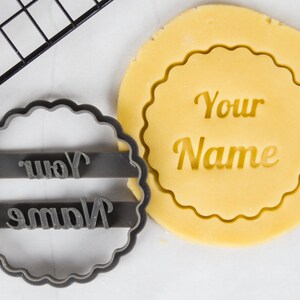 May include: A gray cookie cutter with the words "Your Name" in a script font. A yellow cookie dough is pressed with the cookie cutter, showing the words "Your Name" in the dough.