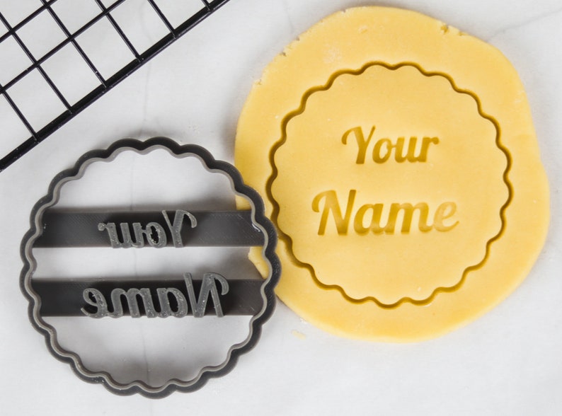 May include: A gray cookie cutter with the words "Your Name" in a script font. A yellow cookie dough is pressed with the cookie cutter, showing the words "Your Name" in the dough.