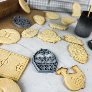 May include: A selection of cookie cutters in various shapes, including a football, a basket, a skull, a flower, and a pair of twins. The cookie cutters are made of metal and have a silver finish.