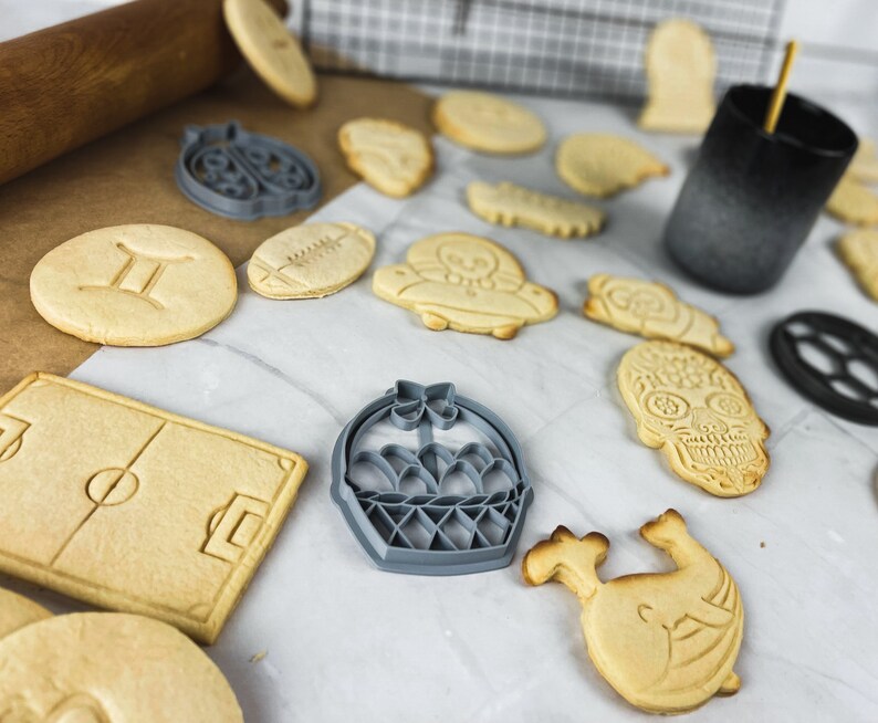 May include: A selection of cookie cutters in various shapes, including a football, a basket, a skull, a flower, and a pair of twins. The cookie cutters are made of metal and have a silver finish.