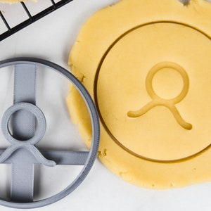 May include: A gray plastic cookie cutter in the shape of the Taurus astrological symbol next to a circle of raw cookie dough with the same design. The dough is light yellow, and the cutter is gray.