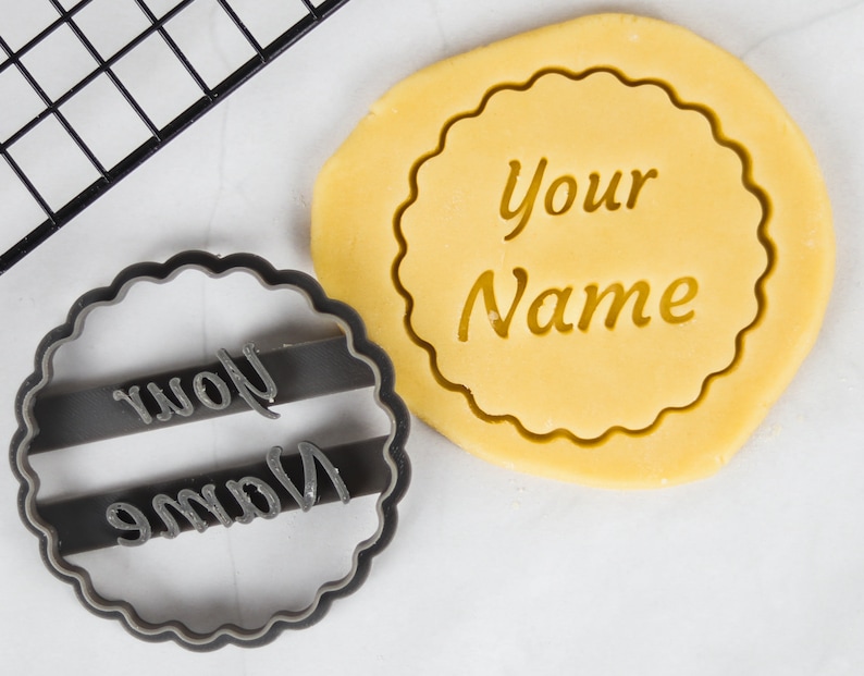 May include: A black plastic cookie cutter with the words "Your Name" in a decorative font. The cutter is on a white surface next to a round piece of cookie dough with the same words imprinted on it.