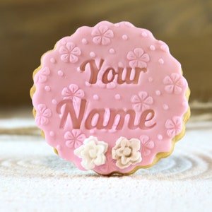 May include: Pink sugar cookie decorated with white flowers and the words "Your Name" in a cursive font.