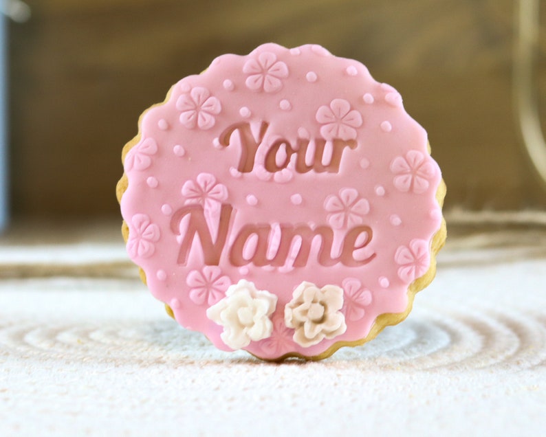 May include: Pink sugar cookie decorated with white flowers and the words "Your Name" in a cursive font.