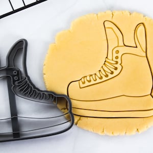 May include: A black plastic cookie cutter in the shape of an ice skate. The cutter is on top of a sheet of unbaked cookie dough.
