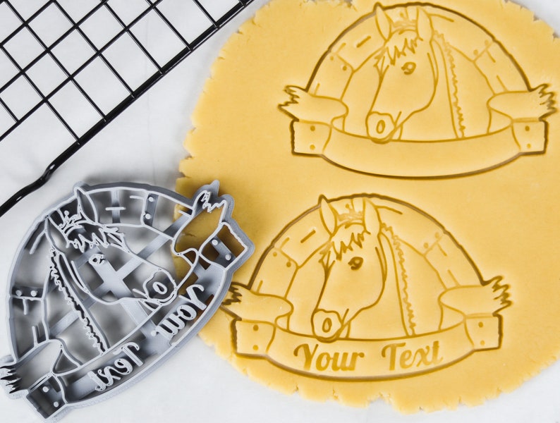 Puede incluir: Un cortador de galletas de pl&aacute;stico gris en forma de cabeza de caballo con una pancarta que dice "Your Text". El cortador de galletas est&aacute; sobre una superficie blanca junto a una masa de galletas amarilla con el mismo dise&ntilde;o estampado.