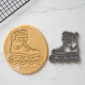 May include: A roller skate cookie cutter next to a cookie with a roller skate imprint. The cutter is gray plastic, and the cookie is round and light brown. A black cooling rack is in the background.