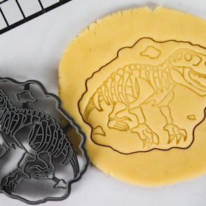 Puede incluir: Un cortador de galletas de plástico negro con forma de esqueleto de dinosaurio. El cortador de galletas está sobre una superficie blanca junto a una pieza redonda de masa para galletas con el diseño del esqueleto de dinosaurio cortado.