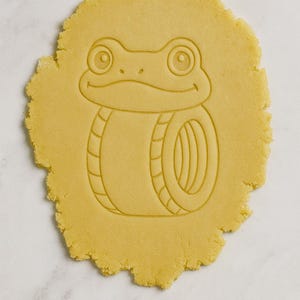 Könnte beinhalten: Ein runder, ungebackener Keksteig-Ausstecher in Form eines Cartoon-Frosches. Der Teig ist hellgelb, und das Froschdesign ist in den Teig geätzt. Der Frosch hat große Augen und einen lächelnden Mund. Für das Backen.