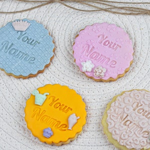 May include: Four decorated sugar cookies with the text "Your Name" on each. The cookies are decorated with different patterns and colors, including blue, pink, yellow, and beige.