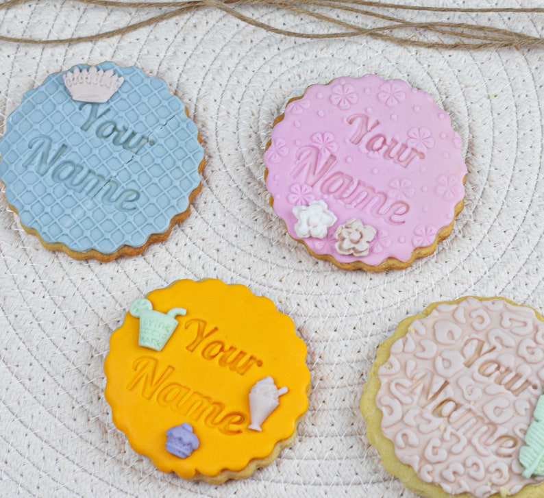 May include: Four decorated sugar cookies with the text "Your Name" on each. The cookies are decorated with different patterns and colors, including blue, pink, yellow, and beige.