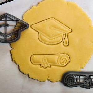 Könnte beinhalten: Ein Ausstechersatz zum Thema Abschluss. Enthält einen Mortarboard, ein Diplom und zwei Ausstecher. Der Mortarboard und das Diplom sind in einen Kreis aus gelbem Teig gedrückt. Die Ausstecher sind grau.