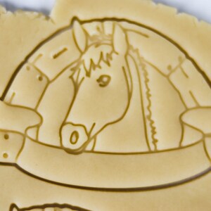 Puede incluir: Un cortador de galletas con forma de cabeza de caballo con una pancarta debajo. El caballo mira hacia la derecha y tiene una crin y una brida. La pancarta tiene dos botones a cada lado.