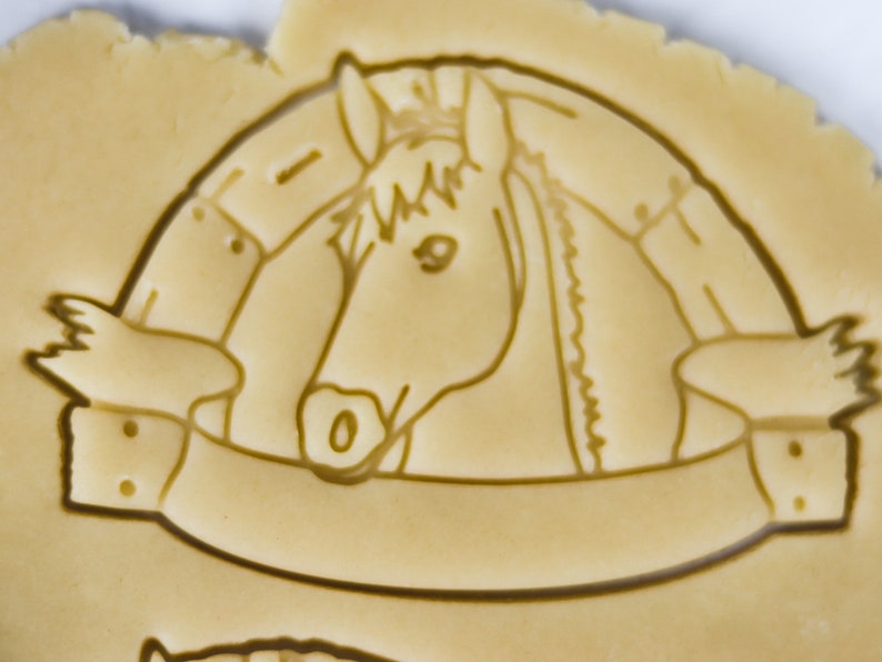 Puede incluir: Un cortador de galletas con forma de cabeza de caballo con una pancarta debajo. El caballo mira hacia la derecha y tiene una crin y una brida. La pancarta tiene dos botones a cada lado.