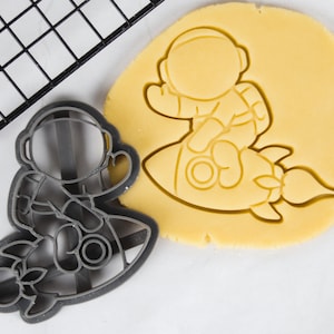 Puede incluir: Un cortador de galletas de plástico gris en forma de astronauta montando un cohete. El cortador de galletas está sobre una superficie blanca junto a una pieza redonda de masa para galletas con el mismo diseño.