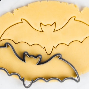 Puede incluir: Un primer plano de un cortador de galletas con forma de murciélago sobre una hoja de masa de galletas amarilla. El cortador de galletas está hecho de metal y tiene un acabado brillante.