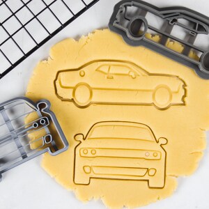 May include: Three cookie cutters in the shape of a car. The cutters are made of gray plastic and are on a white surface. The cutters are next to a sheet of yellow cookie dough.